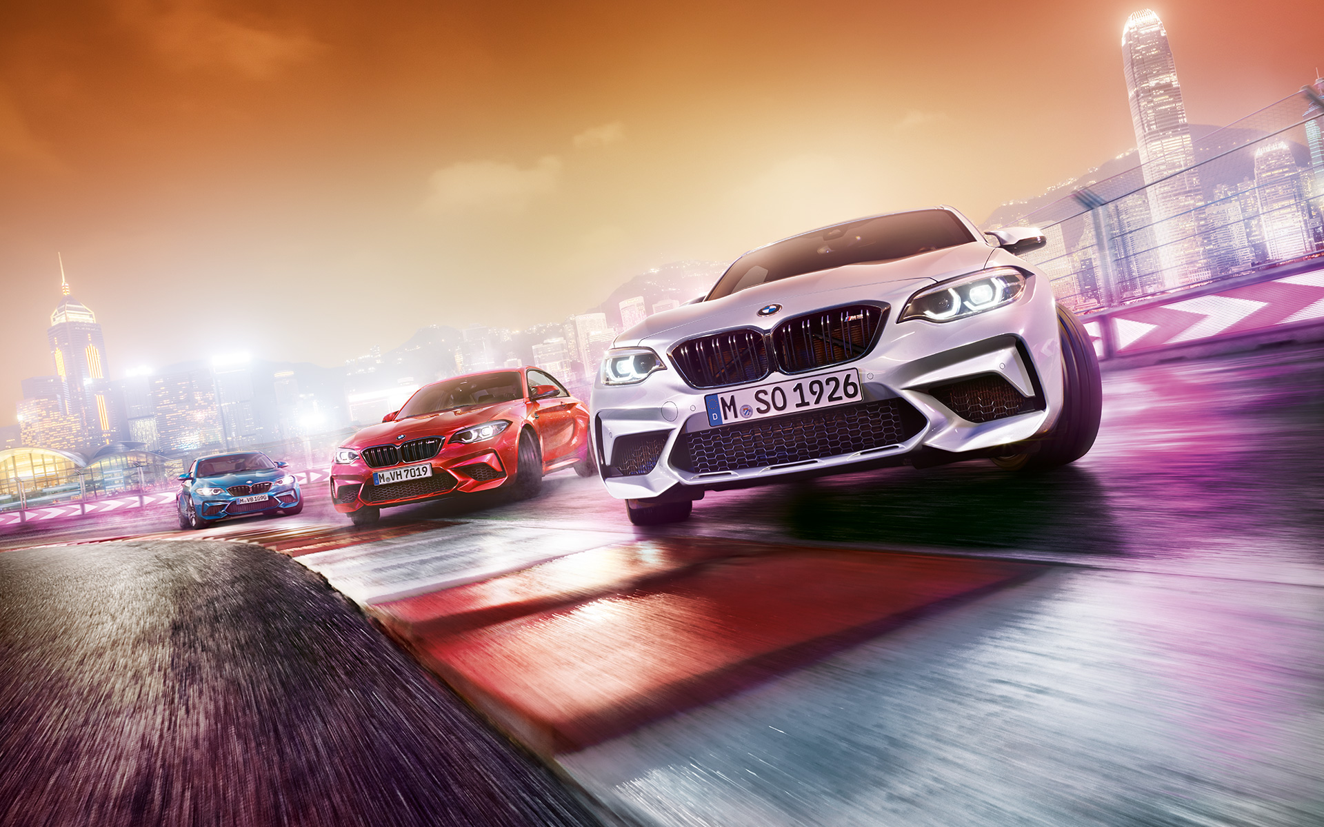 BMW M2 Competition en action Trois BMW M2 Competition font des dérapages sur un circuit de course, dans le décor nocturne d'une grande ville.