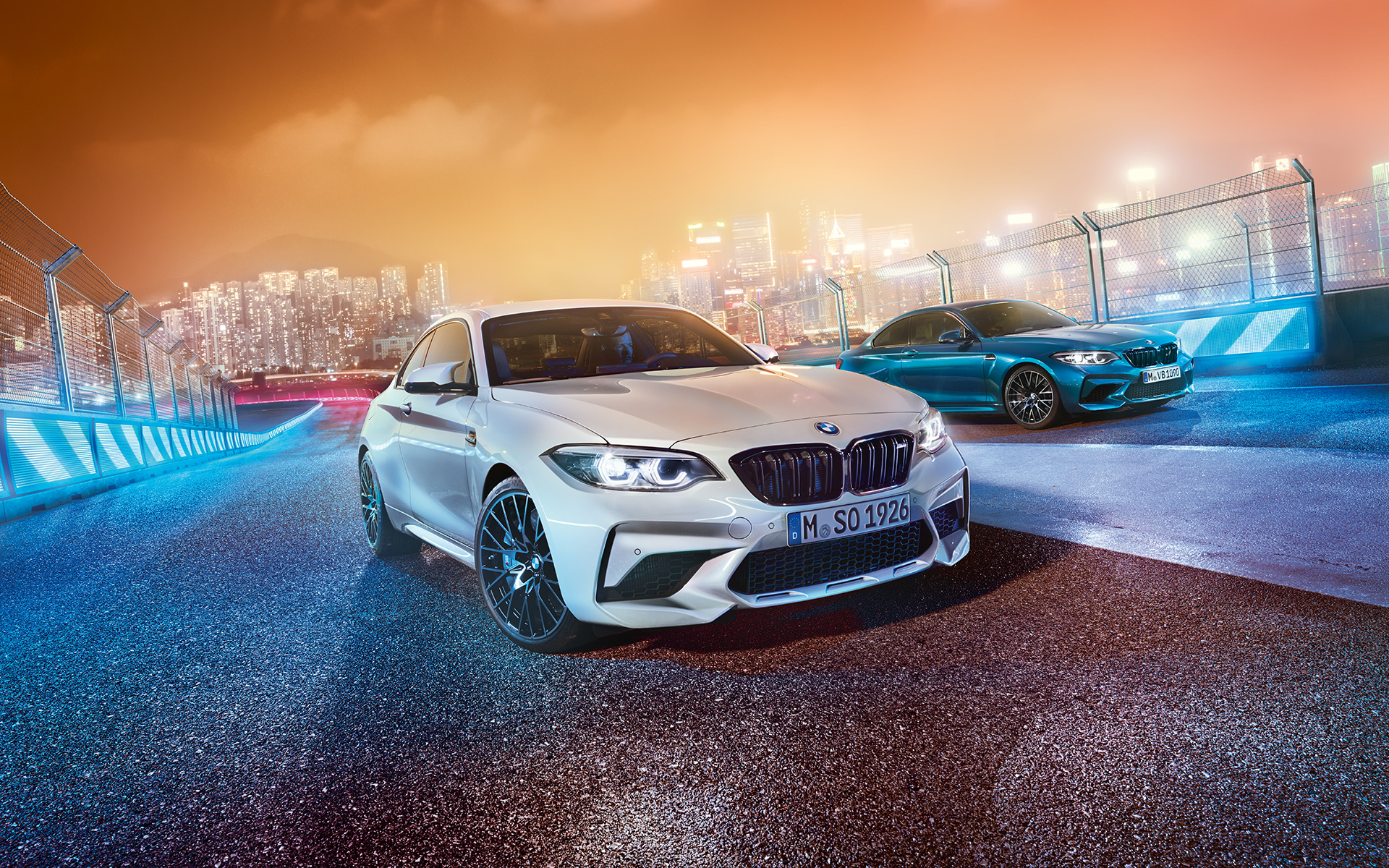 Deux BMW M2 Competition sur le circuit de course. Une BMW M2 Competition blanche et une autre bleue, vue oblique de l'avant, dans le décor nocturne d'une grande ville.