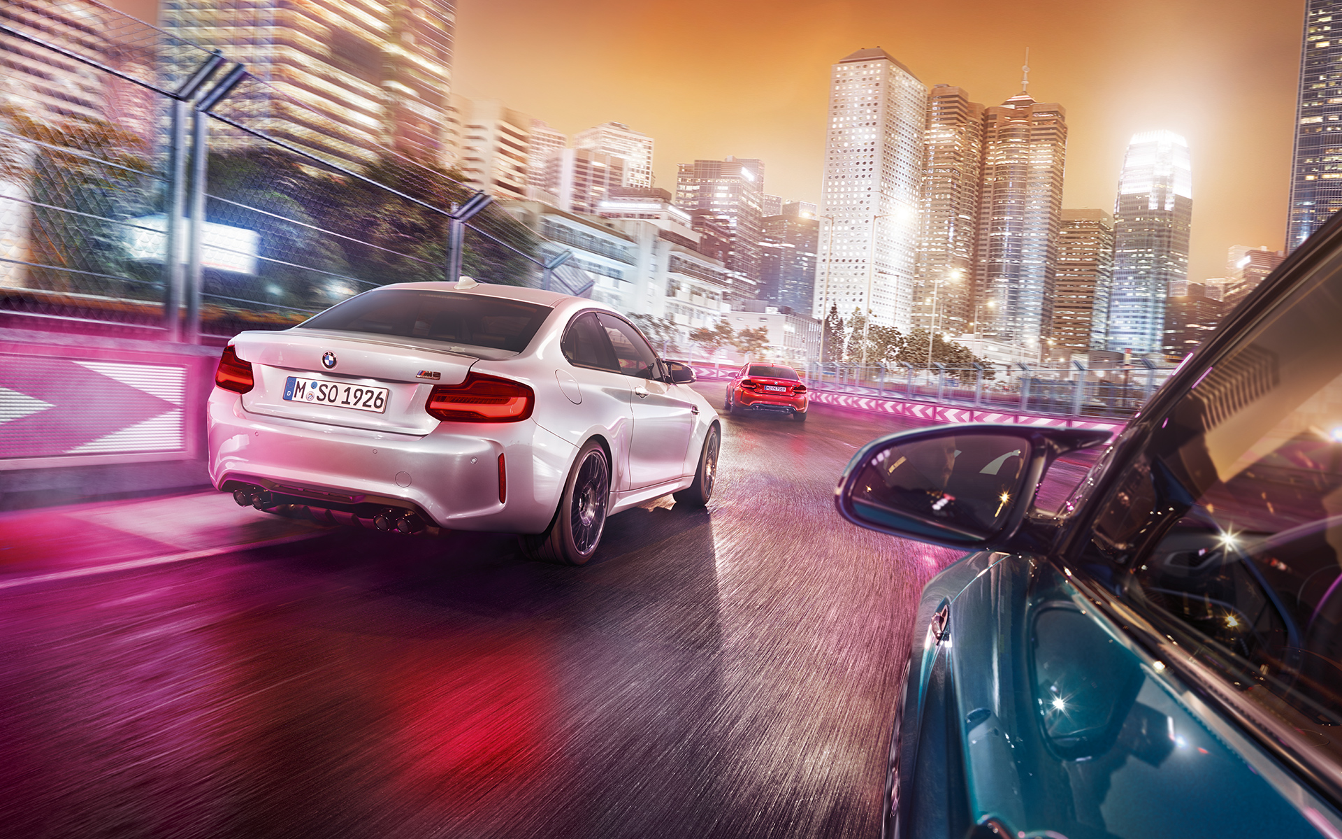 Vue arrière de la BMW M2 Competition Trois BMW M2 Competition vues de l'arrière roulent sur un circuit de course, devant une skyline éclairée.