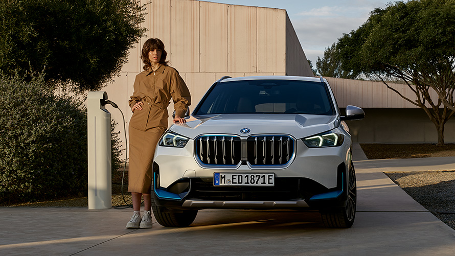 Homme vêtu d’un manteau bleu pétrole devant une BMW iX3 G08 2021 Homme vêtu d’un manteau bleu pétrole devant une BMW iX3 G08 2021 en cours de recharge dans un garage – vue de côté